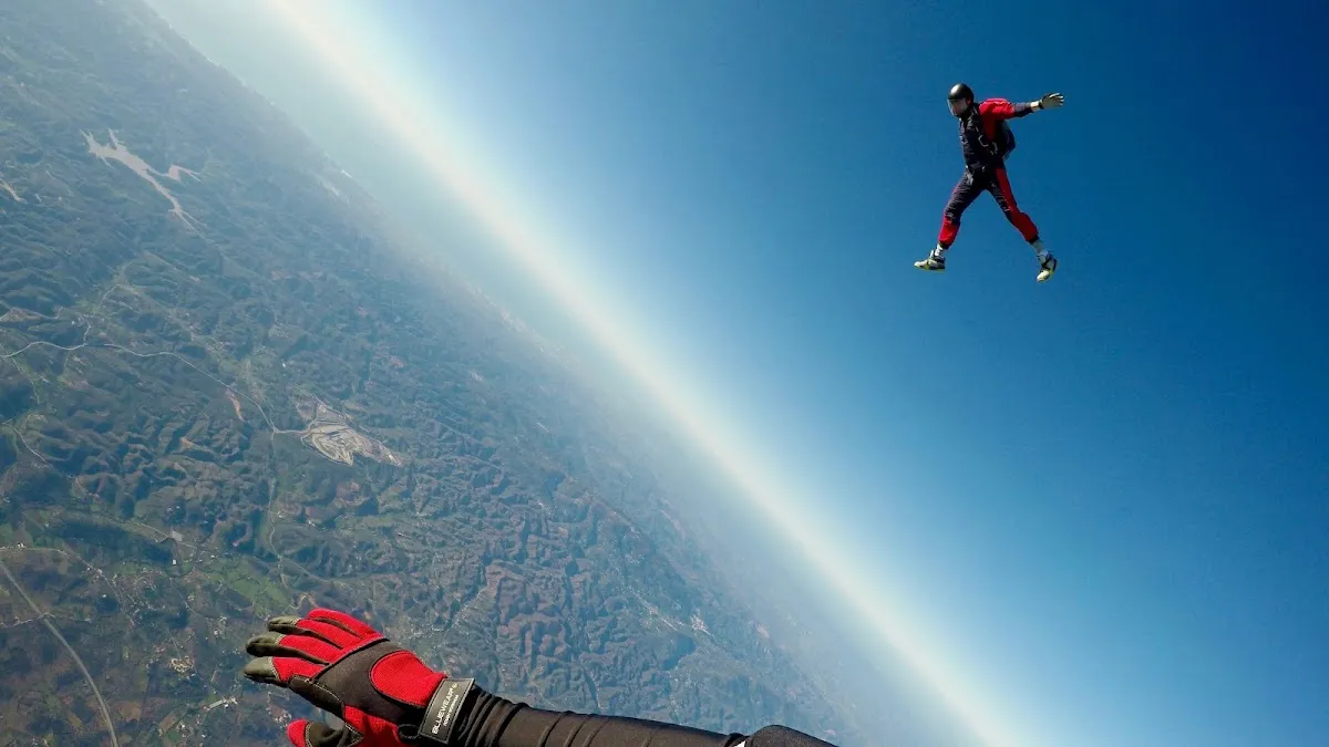 A skydiver in a red suit freefalling high above the earth, photographed from another skydiver reaching out a gloved hand
