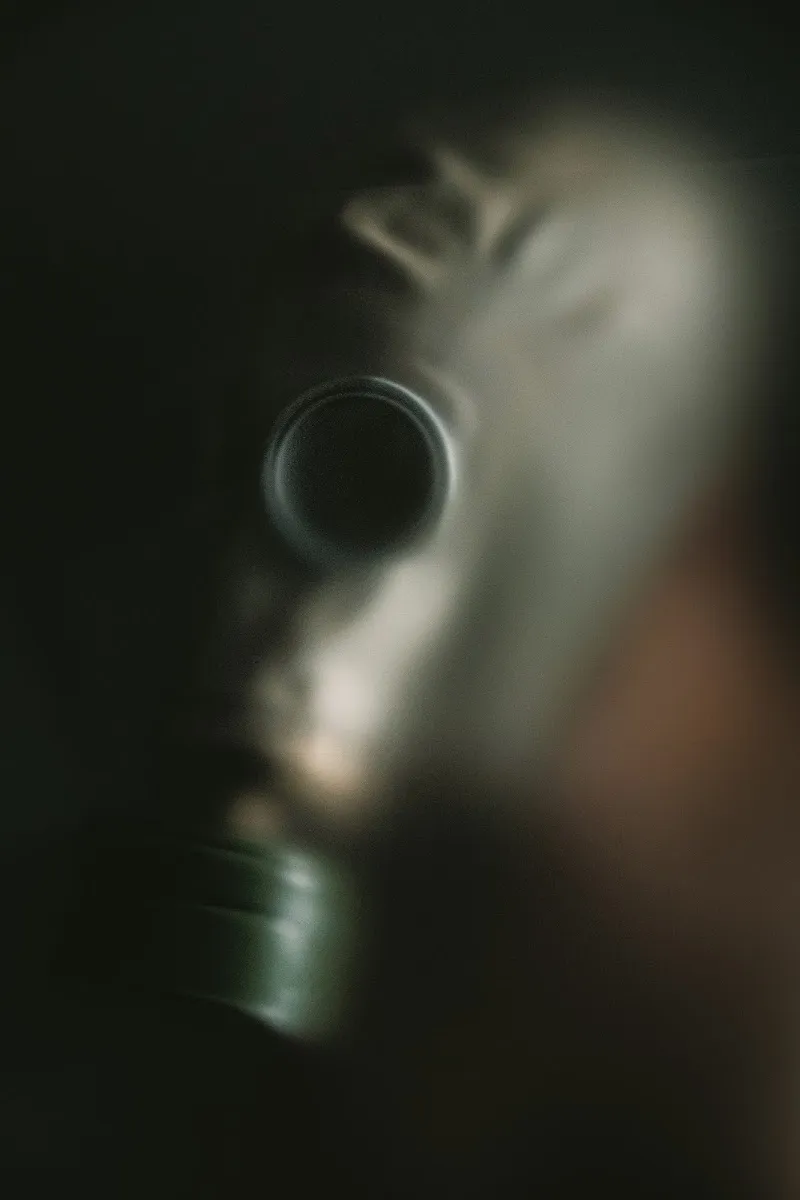 Blurred close-up photograph of a gas mask emerging from darkness, evoking hidden identity