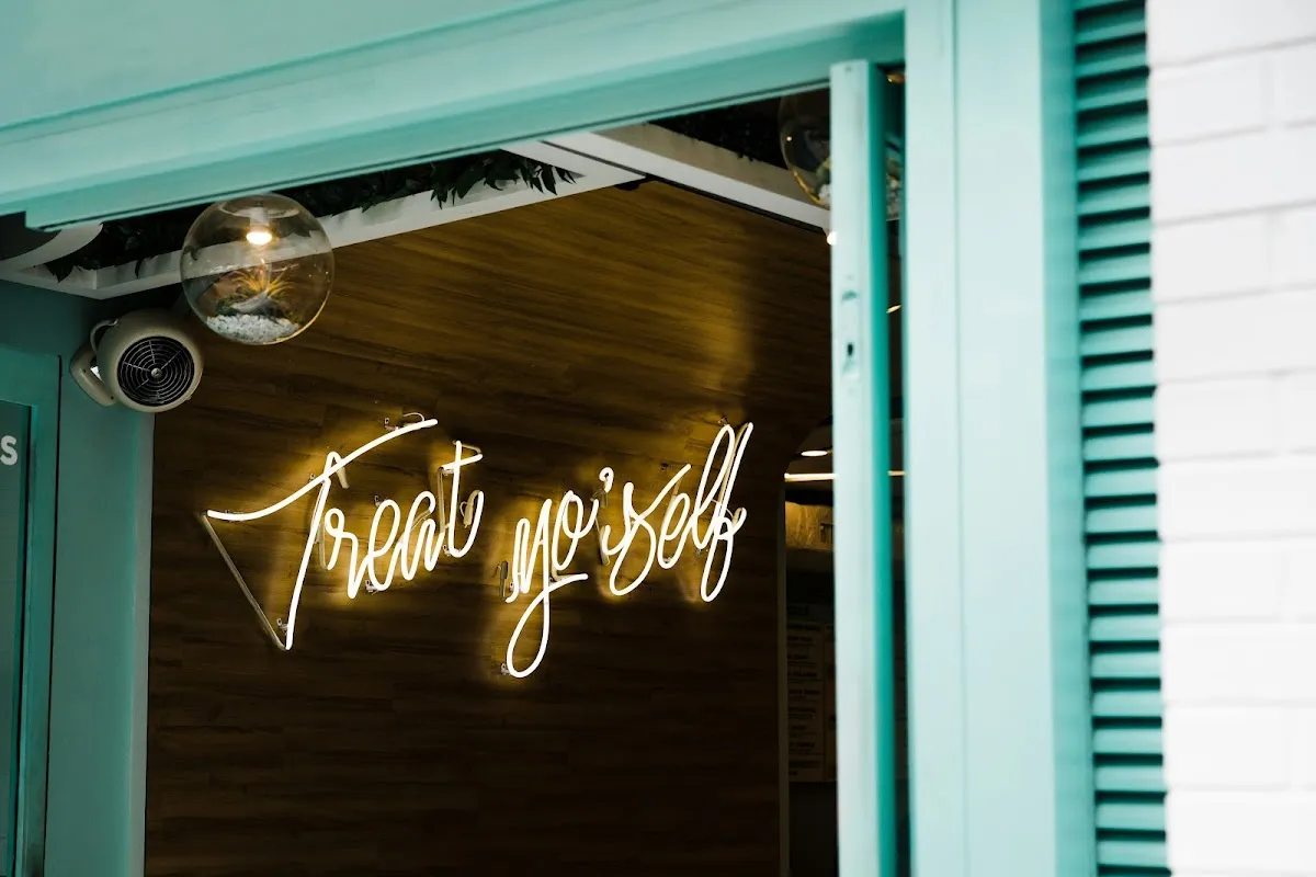 A neon sign reading Treat Yo Self in cursive white light, glowing in a shop window with a turquoise frame