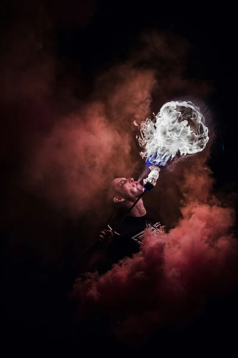 A fire breather performing at night, a large ball of white flame erupting from their mouth amid swirling red and dark smoke