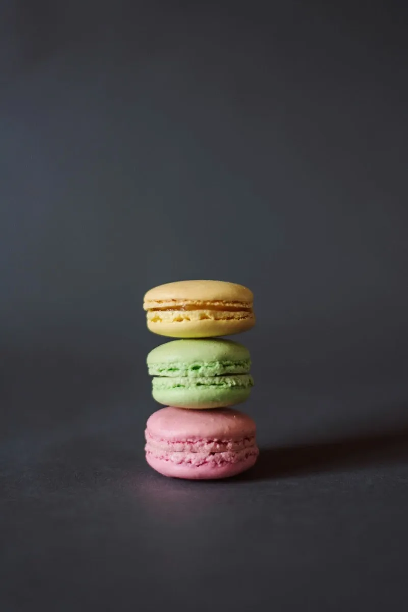 Three pastel macarons — yellow, green, and pink — stacked on top of each other against a dark grey background