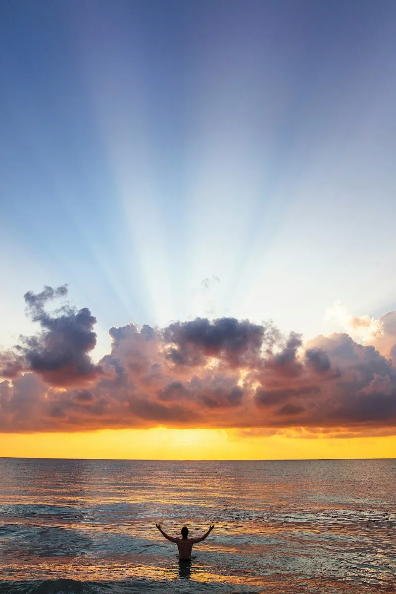 Silhouette of a person standing in the ocean at sunset with arms raised, sun rays bursting through dramatic clouds