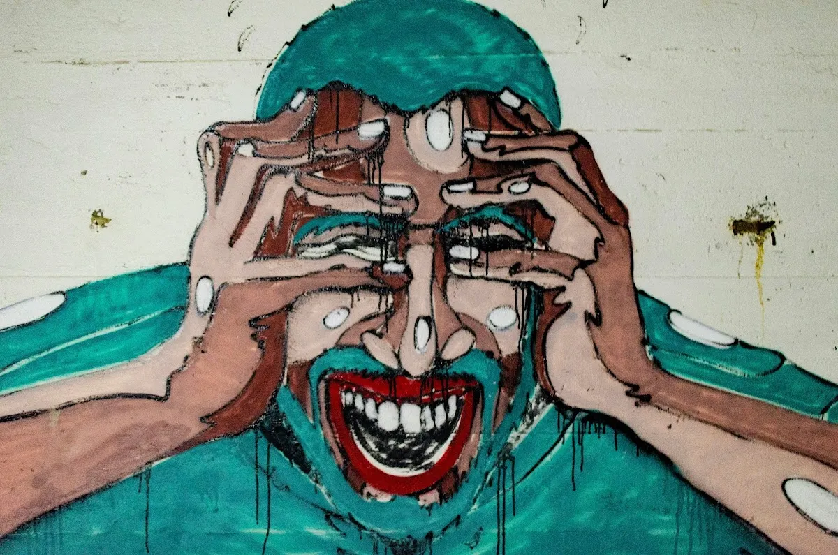 Street art mural of a person in teal clothing gripping their face with both hands, mouth wide open in a scream of overwhelm